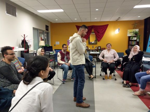 One student stands in the middle of the room thinking surrounded by a group of students sitting in chairs in a circle fashio