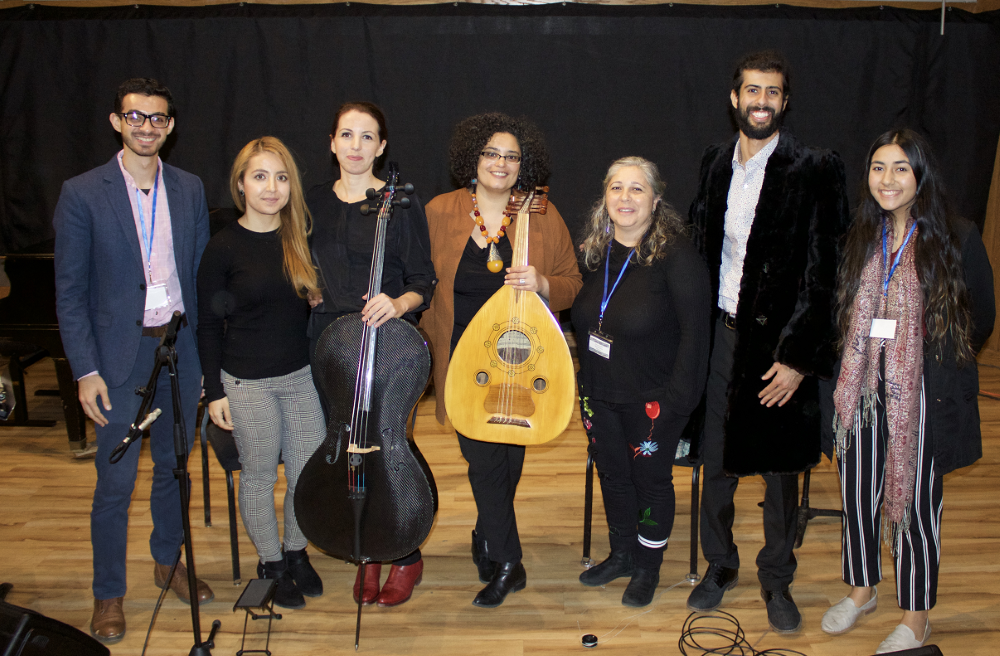 Seven people stand posing for a picture. The third from the left is holding a Cello and the one next is holding a oud.