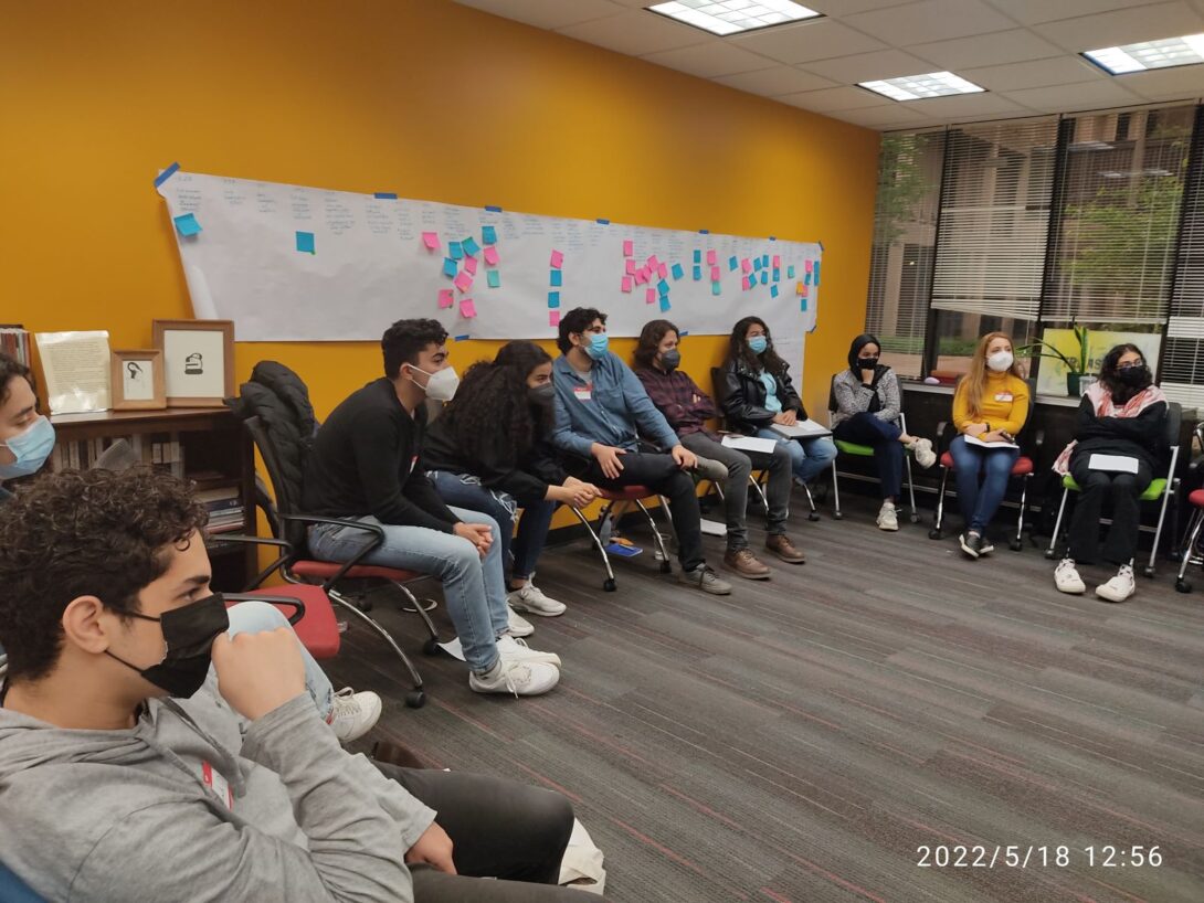 Students sitting in chairs in a semi open circle with masks listening to a presentation. A yellow wall with a white butcher paper with blue and pink sticky notes is behind them