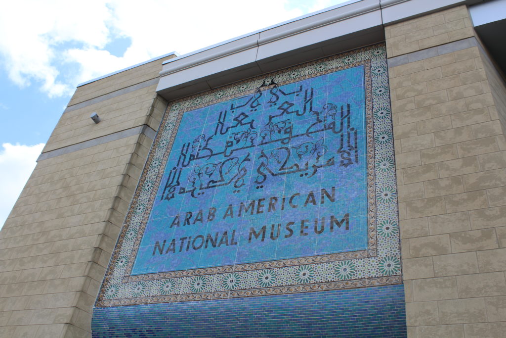 a photograph of a building facade with blue sky with white clouds. Building is beige with a middle section in blue tile with arabic and english calligraphy of the name of the museum