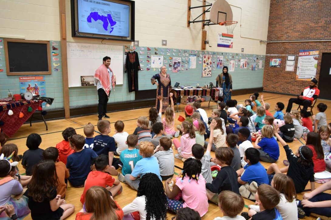 A photograph of elemenatry school children sitting on the floor of the school gym with three adults standing in front of them presenting and a screen on the wall that has the power point presentation.