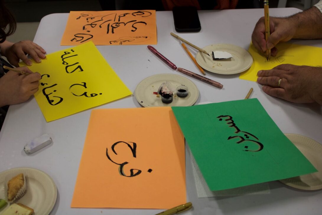 colorful card stock paper with Arabic words in calligraphy on a table with two sets of hands writing on each side of the table.