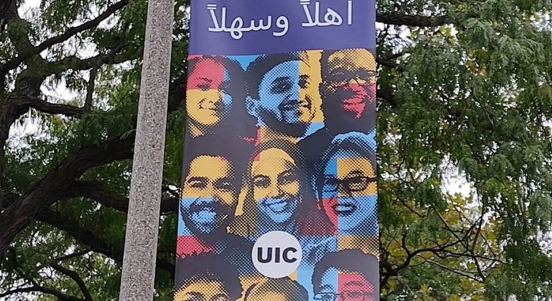 A banner on a pole with trees in the background at UIC campus. The Banner is a UIC banner with images of different students painted in blue, red, yellow, and light yellow with the word Welcome in Arabic
